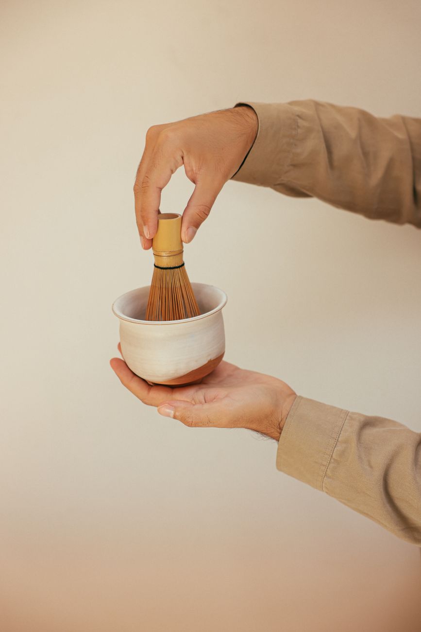 Bamboo chasen whisk creating foam in a ceramic bowl filled with vibrant green matcha tea on wooden surface