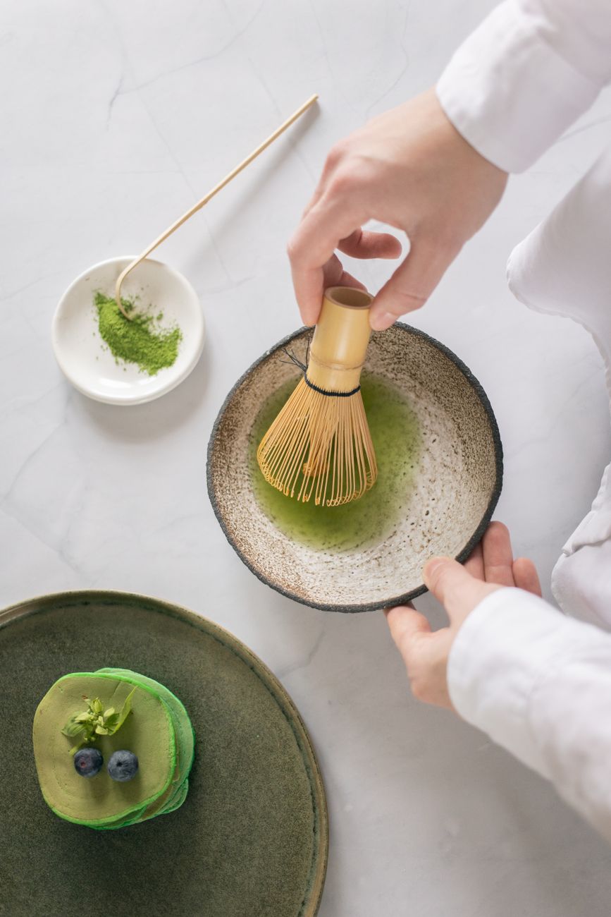 Bamboo chasen whisking vibrant green matcha powder in a ceramic bowl, creating smooth foam with traditional W-shaped hand motions.
