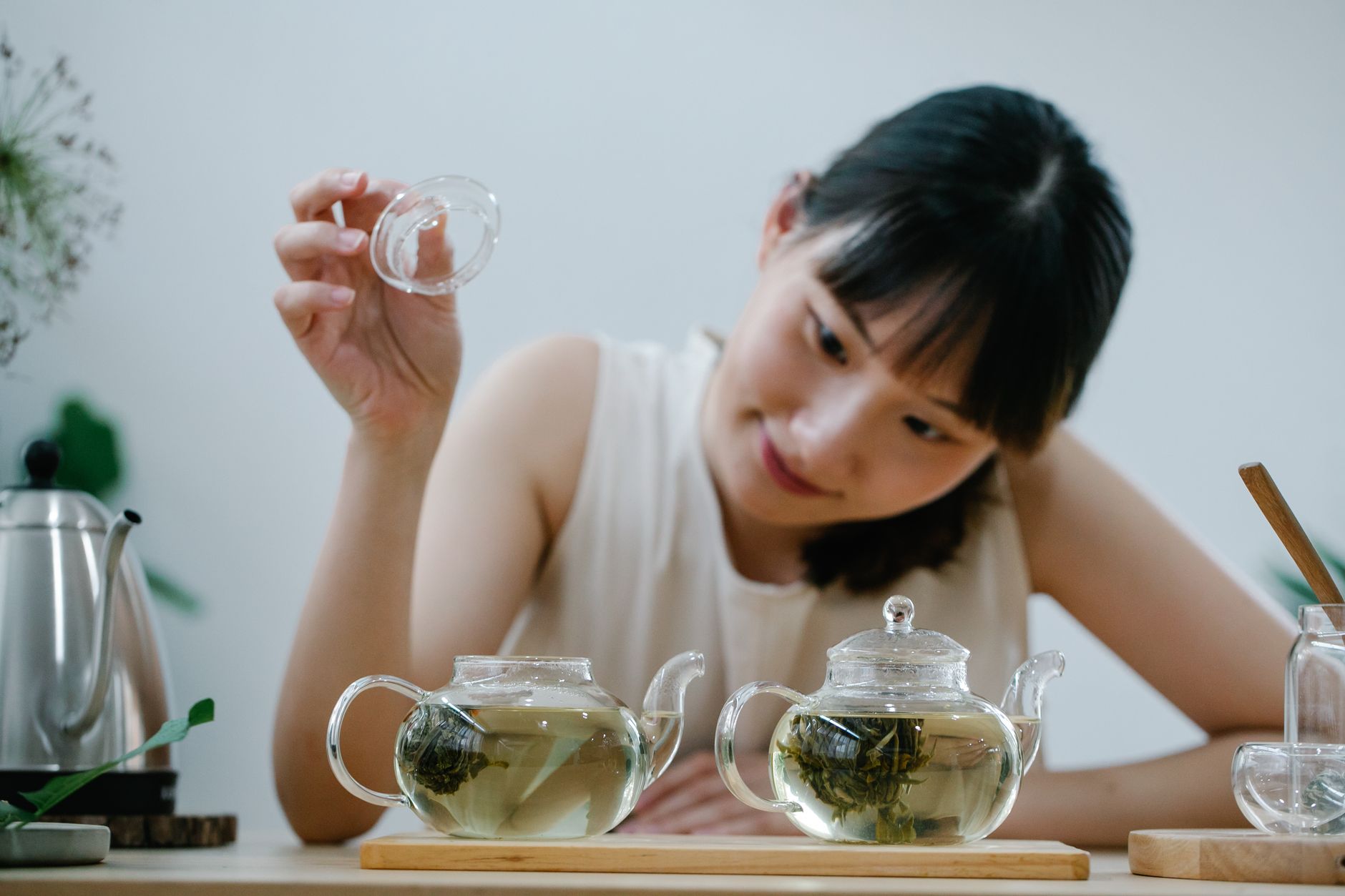 Glass pitcher filled with pale green cold brew tea beside fresh sencha leaves on a wooden table