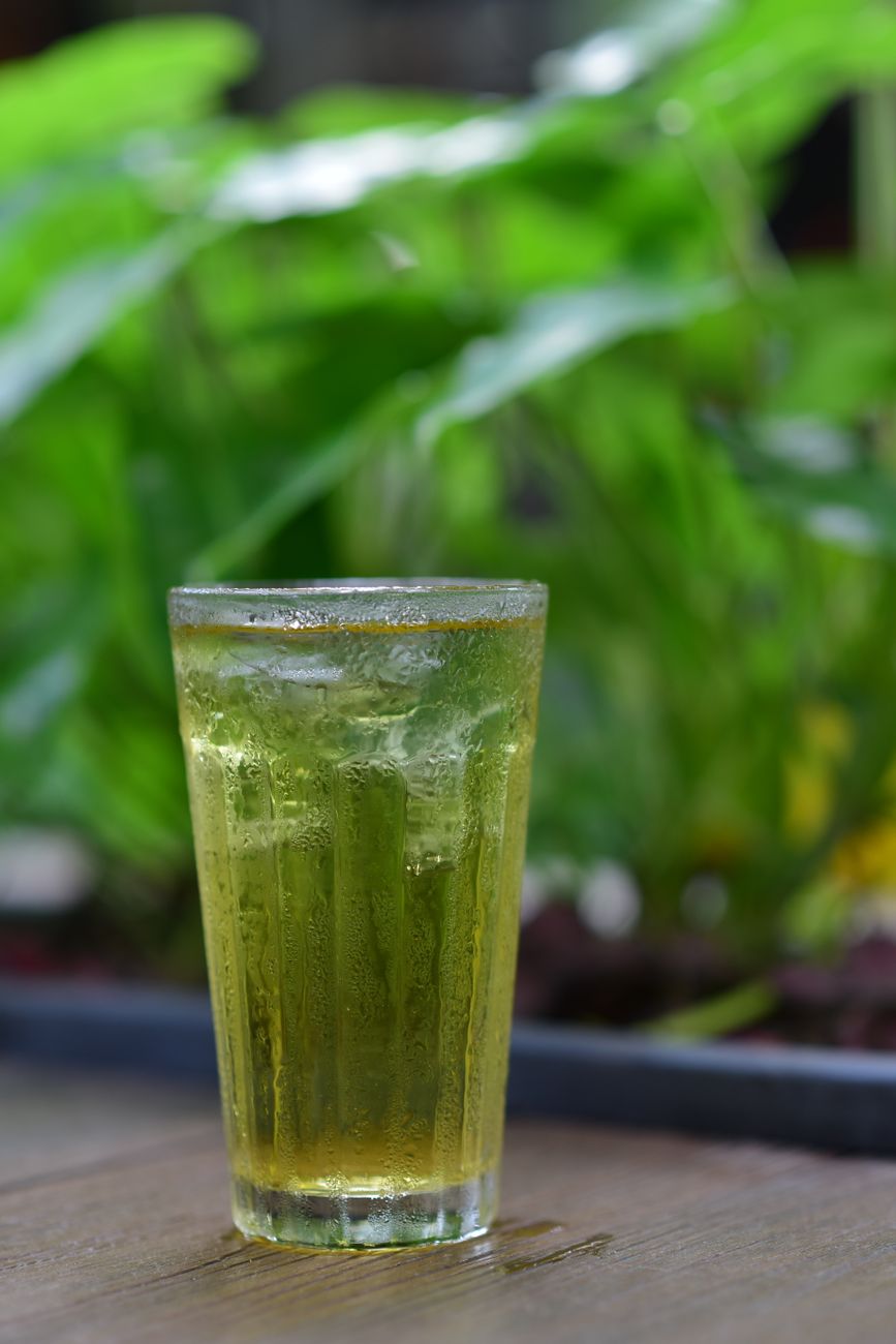 Glass pitcher filled with pale green cold brew tea beside fresh sencha leaves on a wooden table