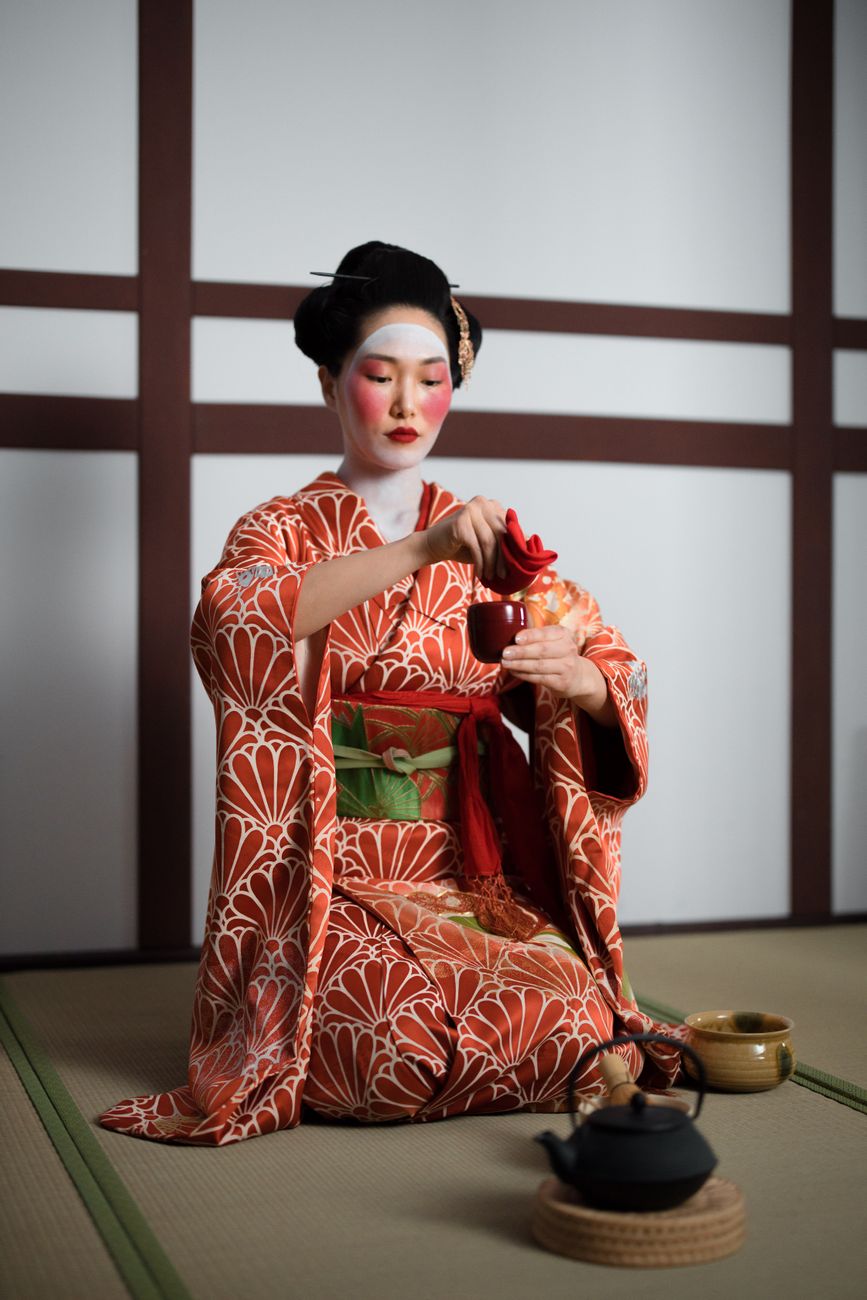 Guest in kimono bowing deeply on tatami mat before entering traditional Japanese tea room with sliding shoji doors.