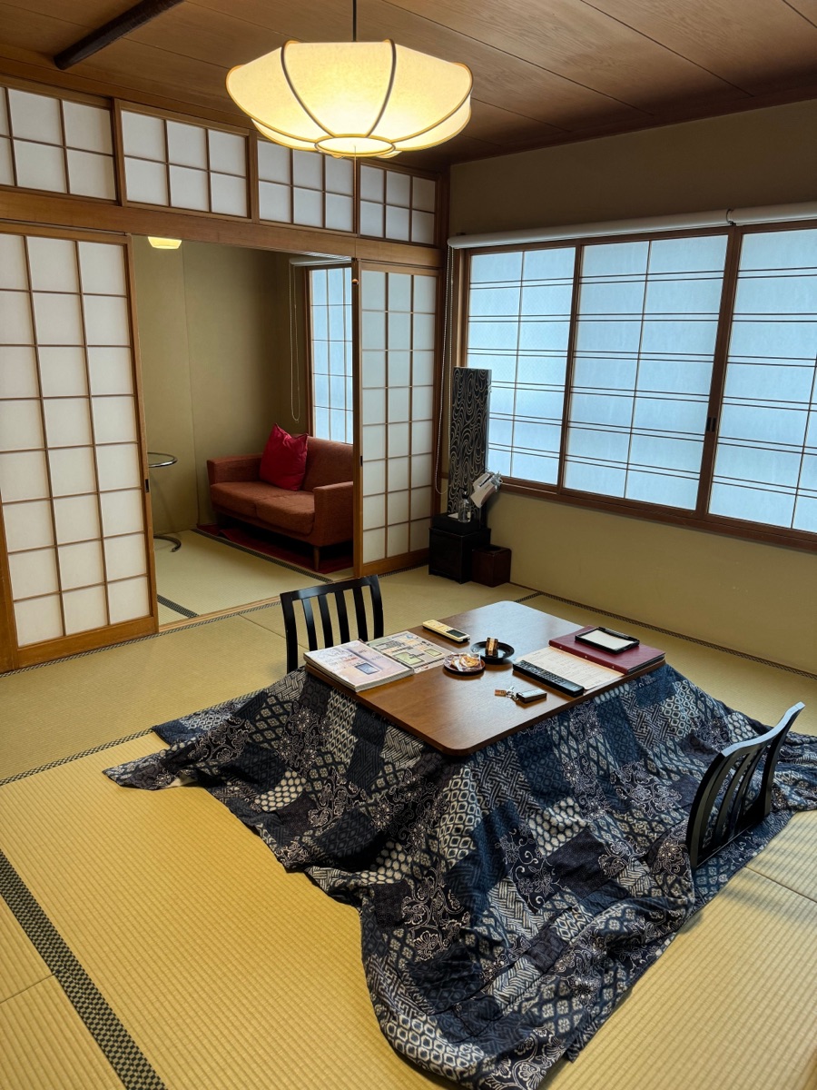 Low wooden kotatsu table with quilted futon draped over frame, steaming tea cup and tangerines on top, warm interior setting