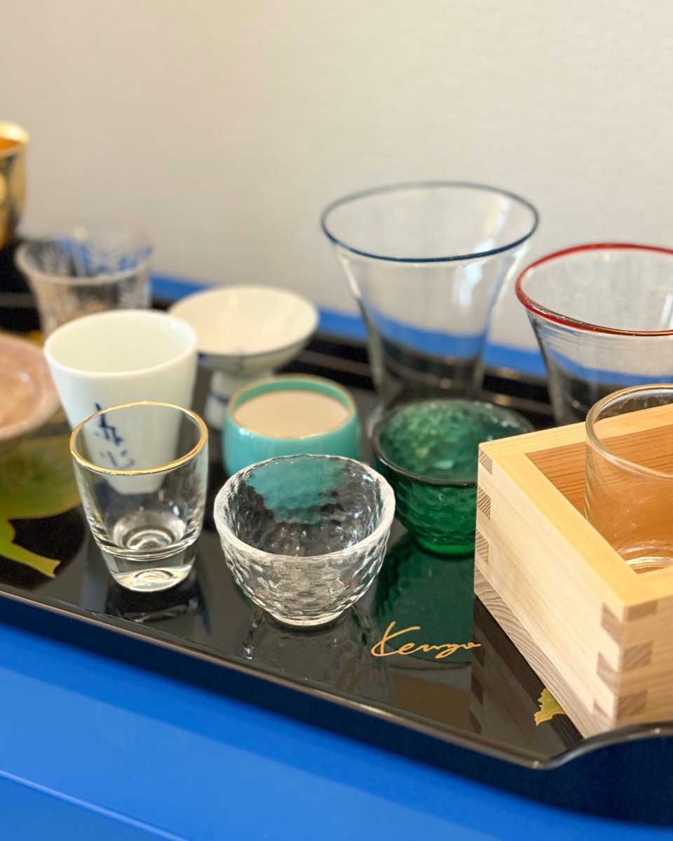 Assorted Japanese sake glasses in varied shapes and materials arranged on a wooden surface with soft natural light