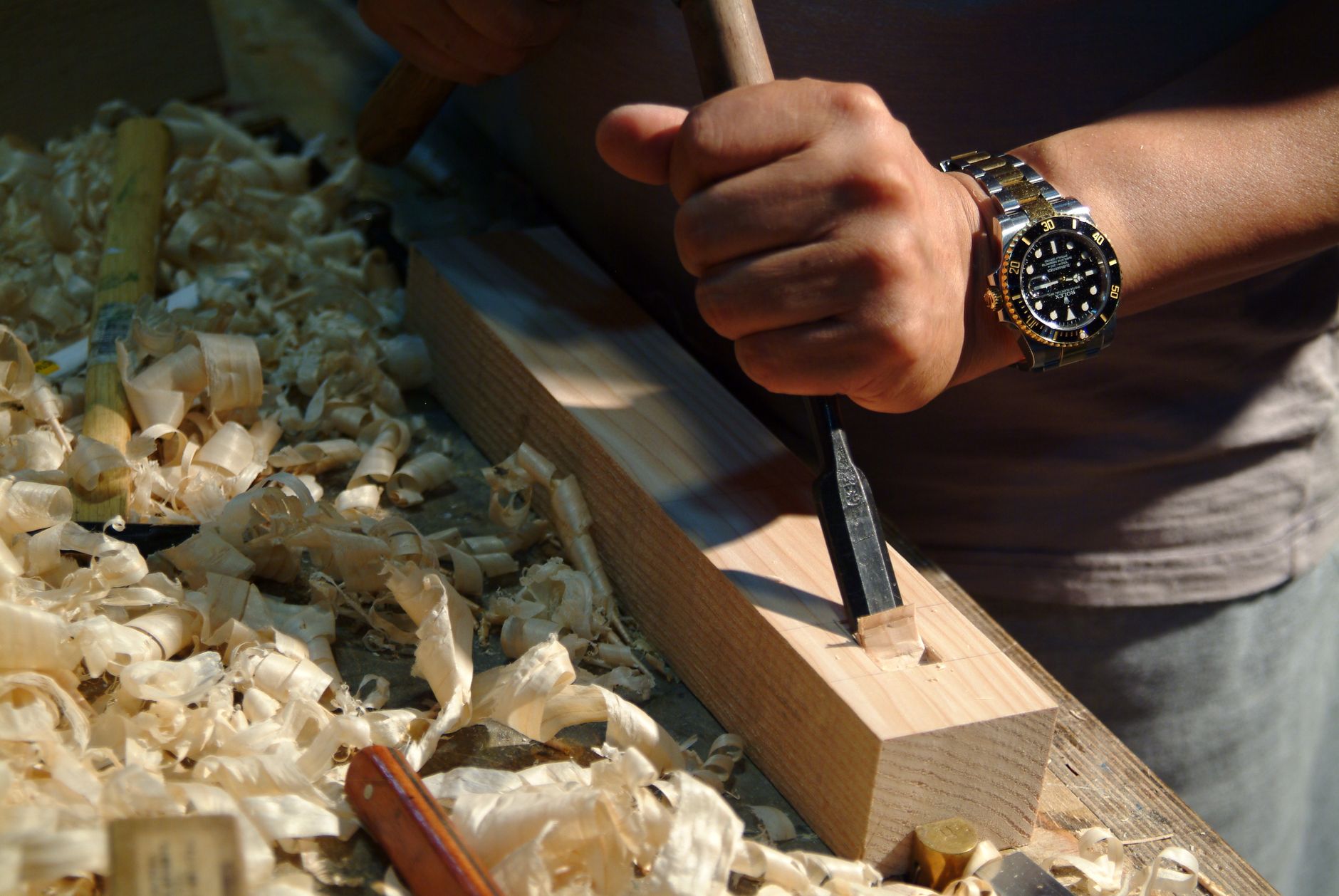 Hands carving intricate details into a wooden block with sharp chisels, wood shavings scattered on a work surface
